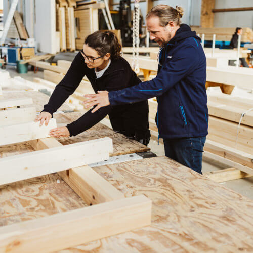 Zimmerer-Team bespricht Holzbauprojekt in Werkstatt, prüft Bauteile und arbeitet gemeinsam an Konstruktion