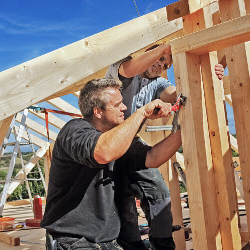 Zimmerer bei Montage einer Holzkonstruktion, arbeiten gemeinsam am Dachstuhl unter blauem Himmel