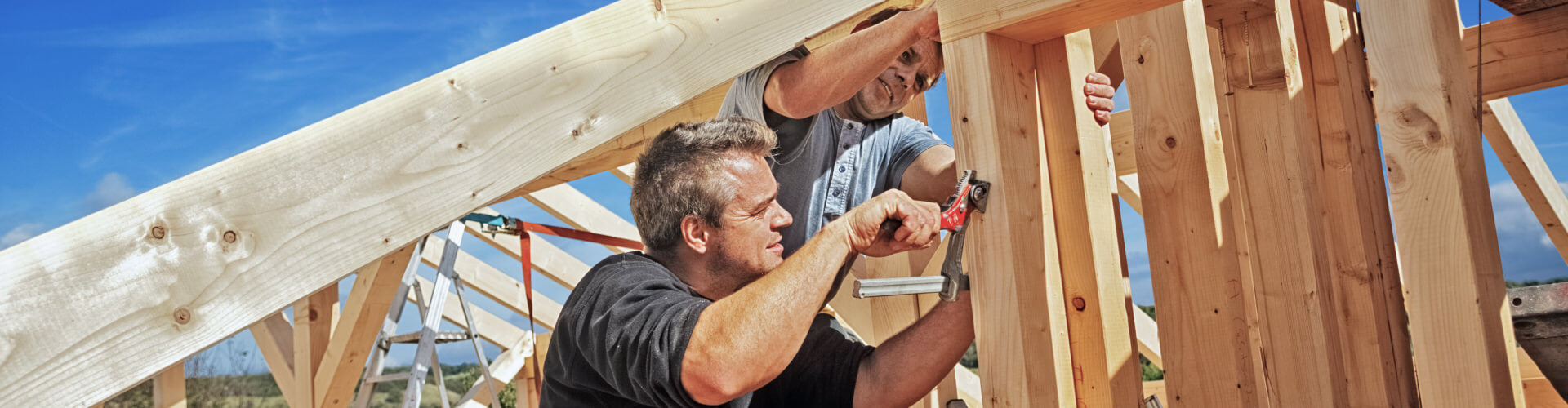 Zimmerer bei Montage einer Holzkonstruktion, arbeiten gemeinsam am Dachstuhl unter blauem Himmel